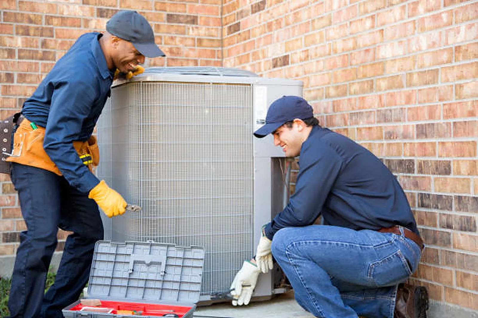 Two HVAC technicians setting an outdoor condenser unit against a brick wall