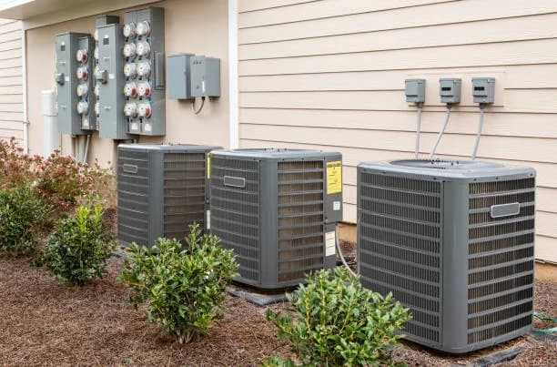 Three central air conditioner condensers on a pad beside a house with landscaping
